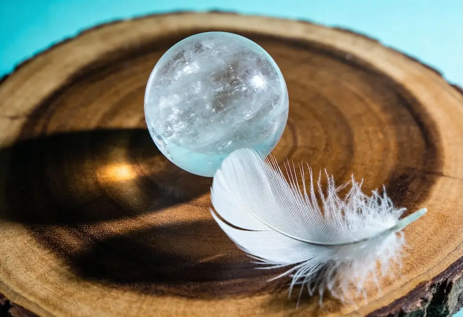 Una esfera de cristal transparente y una pluma blanca descansan sobre una losa redonda de madera con anillos de árboles visibles, sobre un fondo azul claro.